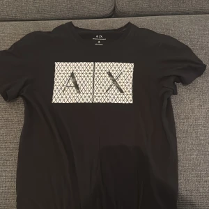 Svart t-shirt från Armani Exchange - Säljer en snygg svart t-shirt från Armani Exchange i storlek S. T-shirten har ett coolt geometriskt mönster på framsidan med två vita rutor och svarta linjer. Den är gjord av bomull och har en slim passform. Perfekt för både vardag och fest!