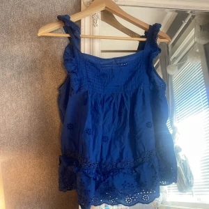 Blå ärmlös topp från MNG - Säljer en superfin blå ärmlös topp från MNG. Den har ett vackert broderat mönster och volangdetaljer vid axelbanden och nederkanten. Perfekt för sommaren och kan stylas både till vardags och fest. Toppen är i bomull och känns väldigt luftig och skön att ha på sig. Passar perfekt till ett par jeans eller en kjol!