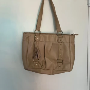 Beige axelväska med fransdetalj - Säljer en snygg beige axelväska i syntetmaterial. Väskan har två handtag och en fransdetalj på framsidan. Den stängs med dragkedja och har flera fack inuti för bra förvaring. Perfekt för vardagsbruk eller en dag på stan!