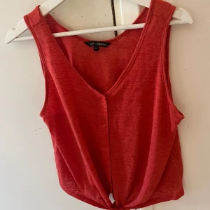 Röd ärmlös topp från KappAhl - Säljer en snygg röd ärmlös topp från KappAhl. Toppen har en v-ringning och knappar framtill. Den är tillverkad i ett mjukt och bekvämt material, perfekt för varma sommardagar. Toppen har också en knytning längst ner för en extra detalj.