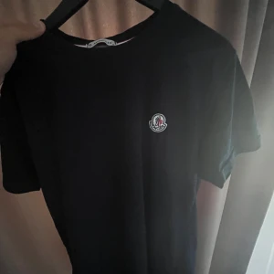 Svart t-shirt från Moncler - Säljer en snygg svart t-shirt från Moncler i storlek M. T-shirten är gjord av 100% bomull och har en liten broderad Moncler-logga på bröstet. Perfekt för både vardag och fest! Den är i nyskick och har aldrig använts. Passa på att köpa denna stilrena t-shirt!