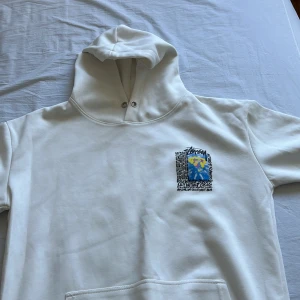 Stussy hoodie - Säljer en vit hoodie från Stüssy med en cool grafisk design på både fram- och baksidan. Den har en stor ficka på magen och en bekväm huva med dragsko. Perfekt för en avslappnad och stilren look! Vid snabb affär 350kr