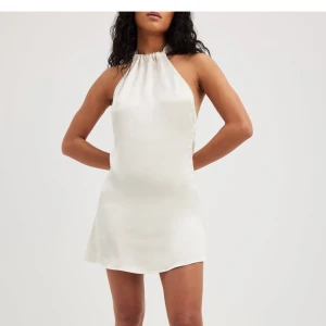 Miniklänning satin med halterneck  - Den här miniklänningen har ett satintyg. Den har en halterneckdesign med en knytbar stängning. Den här miniklänningen kommer i naturvitt.