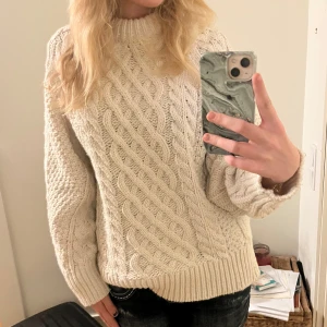 Beige stickad tröja - Säljer en supermysig vit stickad tröja. Den har ett snyggt flätmönster och är perfekt för kalla dagar. Tröjan har långa ärmar och ribbstickade muddar vid ärmslut och nederkant. Perfekt för höst och vinter! 🧡