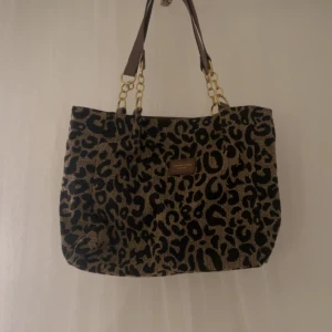 Leopardmönstrad shoppingväska med kedjedetaljer - Säljer en snygg leopardmönstrad shoppingväska med bruna handtag och guldfärgade kedjedetaljer. Väskan är rymlig och perfekt för shopping eller vardagsbruk. Den har en dragkedja för att hålla dina saker säkra. Väskan är i bra skick och redo för en ny ägare!