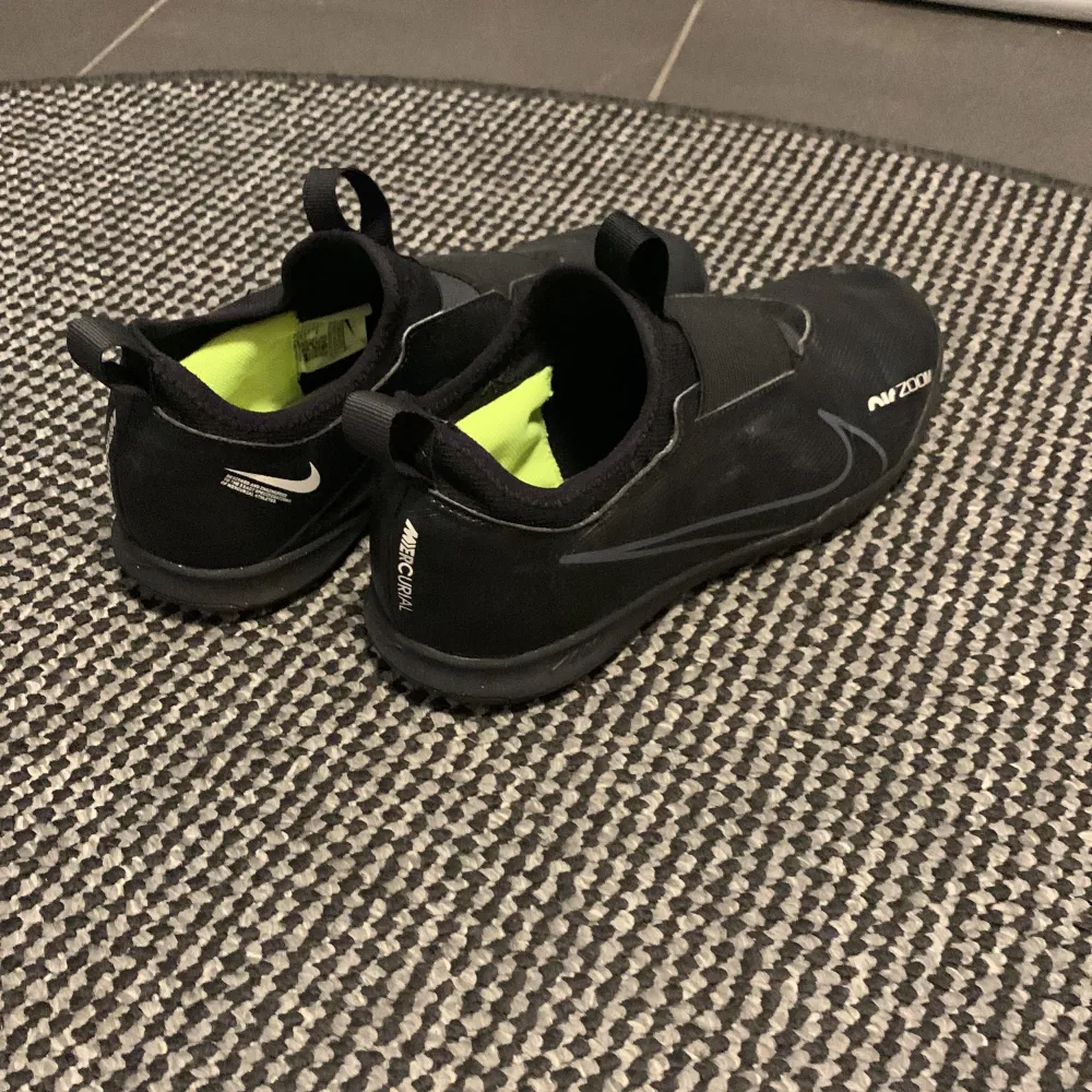 Säljer ett par svarta Nike Air Zoom fotbollsskor i bra skick. Skorna har en modern design med en elastisk rem över vristen för extra stöd och en bekväm passform. Insidan är neonfärgad vilket ger en cool kontrast. Perfekta för fotbollsträning och matcher inomhus. en liten defekt på högra skon . Skor.