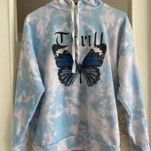 Blå hoodie med fjärilsmotiv - Säljer en superfin blå hoodie med ett stort fjärilsmotiv och texten 'Thrill' på framsidan. Hoodien har en cool tie-dye effekt i blått och vitt, och är perfekt för både höst och vår. Den är i nyskick och har en normal passform. Perfekt för en avslappnad och stilren look!