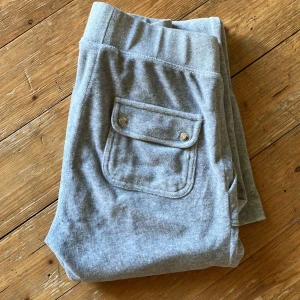 Grå mjukisbyxor från Juicy Couture - Säljer ett par supermysiga grå mjukisbyxor från Juicy Couture i storlek XS. Byxorna har en bekväm passform med elastisk midja och snörning. De har också praktiska fickor med knappar och en snygg broderad logga på ena benet. Perfekta för en chill dag hemma eller en snabb tur till affären. 🩶Ett litet märke på ena smalbenet är enda defekten. skicka dm vid frågor. 🌸