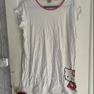 Vit Hello Kitty t-shirt - Säljer en söt vit t-shirt från Hello Kitty i storlek XL. T-shirten har en rosa kant runt halsen och ett stort Hello Kitty-tryck längst ner. Perfekt för en avslappnad dag eller som en mysig pyjamaströja. Den är i bra skick och väldigt bekväm att ha på sig.