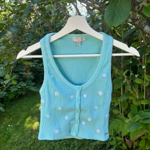 Pacific republic - Pull and bear topp - En turkos kort topp från pacific republican - Pull and bear. På framsidan är det vita och gula broderade blommor samt knappar. Använd 1 gång