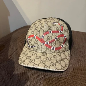 Gucci keps med ormtryck - Säljer en snygg Gucci keps med ett coolt ormtryck på framsidan. Kepsen är beige med det klassiska Gucci-mönstret och har en svart mesh-baksida. Perfekt för att ge din outfit en lyxig touch! (priset går att diskutera)