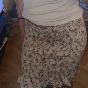 Blommig kjol med volang - Säljer en superfin blommig kjol med volangdetaljer. Kjolen är i en härlig beige färg med små rosa och gröna blommor. Den är perfekt för sommaren och passar både till vardags och fest. Materialet känns som bomull och den har en tight passform som framhäver figuren. Perfekt att matcha med en enkel topp eller blus!