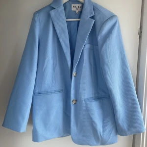 Ljusblå kavaj från NA-KD - Säljer en snygg ljusblå kavaj från NA-KD i storlek 36. Kavajen har en klassisk design med två knappar framtill och två fickor. Den är tillverkad i ett syntetmaterial. Kavajen är i bra skick men har en liten reva på insidan som syns på sista bilden.