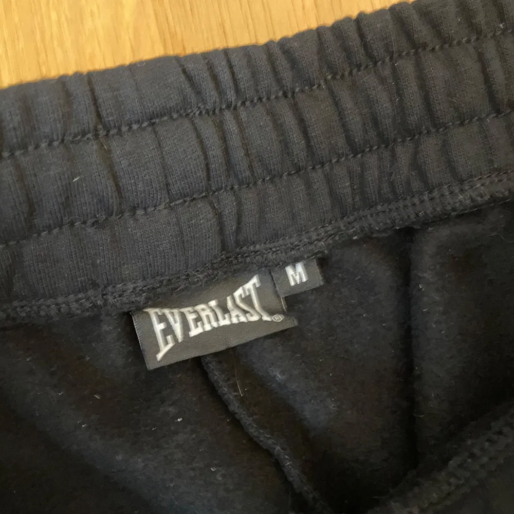 Säljer ett par svarta mjukisbyxor från Everlast i storlek M. Byxorna har en elastisk midja med snörning för bästa passform och är gjorda i ett mjukt och bekvämt material. Perfekta för träning eller en avslappnad dag hemma. Everlast-loggan finns tryckt i vitt längs benet.. Jeans & Byxor.