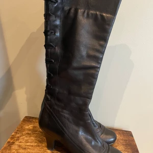 Svarta stövletter med klack - Säljer ett par eleganta svarta stövletter i skinn med klack. De har en snygg snörning på baksidan som ger en unik touch. Perfekta för höst och vinter, och passar både till vardags och fest. Superfin sula och klack. Märke KBM.