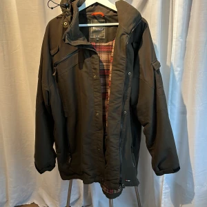 Grön jacka från Pinewood - Säljer en grön jacka från Pinewood i storlek 4XL. Den har en praktisk huva och flera fickor med dragkedja. Insidan är fodrad med ett rutigt mönster i rött och beige. Perfekt för höst- och vinterväder!