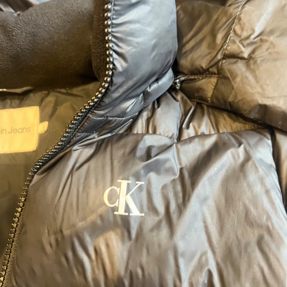 Säljer en snygg svart pufferjacka från Calvin Klein Jeans. Jackan är perfekt för kalla dagar med sin vadderade design och har en praktisk dragkedja framtill. Den har en avtagbar huva och två fickor med dragkedja. Perfekt för både vardag och lite finare tillfällen!. Jackor.