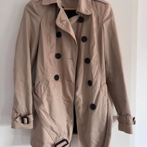 trenchcoat  - ljusbrun trenchcoat med jättefint spänne, perfekt till hösten. köpt secondhand men helt oanvänd av mig. 78 cm från axel till nederkant. Liten i storleken