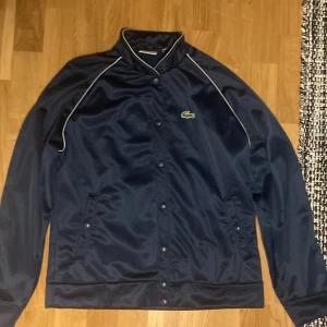 Svart jacka från Lacoste - Säljer en snygg svart jacka från Lacoste med  krokodilloggan broderad stort på ryggen. Jackan har långa ärmar och knappar framtill dock lite sletna (se bild) Men annars är den för en sportig jacka och är väldigt laidback old money.
