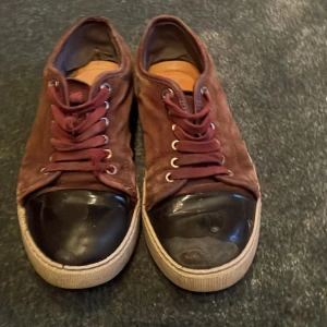 Bruna mockasneakers från Lanvin - Säljer ett par vinröda sneakers från Lanvin i mocka. De har en cool svart tåhätta i skinn och snörning framtill. Sulan är i en ljusare beige ton som ger en snygg kontrast. Perfekta för en avslappnad men stilren look! Sulan är utbytt men medkommer vid