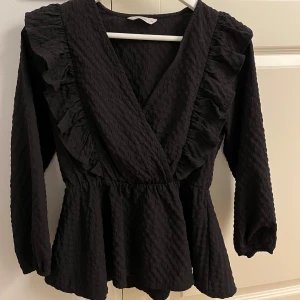 Svart blus med volangdetaljer - Säljer en elegant svart blus med volangdetaljer. Den har en omlott-design och långa ärmar, vilket gör den perfekt för både vardag och fest. Materialet är mjukt och bekvämt, vilket ger en fin passform.