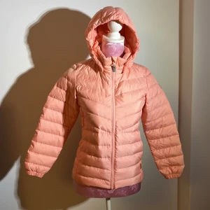 Rosa tunn pufferjacka från Reima - Säljer en superfin rosa pufferjacka från Reima i storlek 140. Jackan är perfekt för kyliga dagar med sitt dun- och fjäderfoder. Den har en dragkedja framtill och en mysig huva. Materialet är 100% polyester, vilket gör den både lätt och varm. Perfekt för höst och vinter!