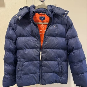 Mörkblå dunjacka från Gant stl 146-152 - Säljer en snygg mörkblå dunjacka från Gant i mycket bra skick. Jackan har en dragkedja framtill och en avtagbar huva. Finns ett litet hål på ärmen som knappt syns.