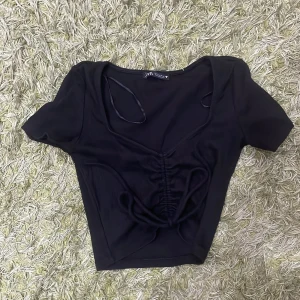 Svart ribbad topp från Zara - Säljer en snygg svart ribbad topp från Zara i storlek S. Toppen har en v-ringad design med knytning framtill som ger en cool look. Perfekt för både vardag och fest! 🖤