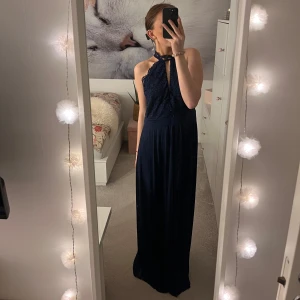 Mörkblå långklänning med spetsdetaljer - Hej! Säljer nu min balklänning som är använd en gång. Det är en elegant mörkblå långklänning med spetsdetaljer och halterneck. Perfekt för festliga tillfällen. Klänningen har en vacker öppen rygg och är ärmlös. I klänningen står att den är i storlek 42 men jag skulle säga att den är en mindre 42 eller en storlek 40. Jag är 173 och den räcker mig ner till marken. 