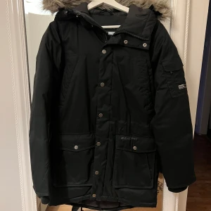 Svart parkajacka med pälsdetaljer - Säljer en svart parkajacka från Everest i mycket bra skick. Jackan har en mysig huva med pälsdetaljer och praktiska fickor med knappar. Perfekt för kalla vinterdagar och höstpromenader. Den är långärmad och har en normal passform. Supervarm och stilren! Stor i storlek, passar mig som är normalt 36/38