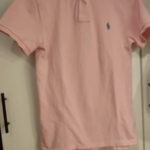 Rosa pikétröja från Ralph Lauren - Säljer en snygg rosa pikétröja från Ralph Lauren i slim fit. Den har korta ärmar och en klassisk krage med knappar. Det ikoniska blå broderade logotypen syns på bröstet. Perfekt för en stilren och avslappnad look! I storlek S