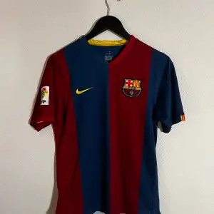 Säljer en FC Barcelona fotbollströja från 2006/07 i klassiska rödblå färger. Tröjan har korta ärmar och klubbens emblem på bröstet. Perfekt för fans som vill visa sitt stöd! 🏆