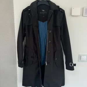 Trenchcoat - Snygg svart trenchcoat från Snoot i storlek XS. Jackan har ett bälte i midjan för en justerbar passform. Den är långärmad med knappar och dragkedja framtill. Perfekt för höst och vår!