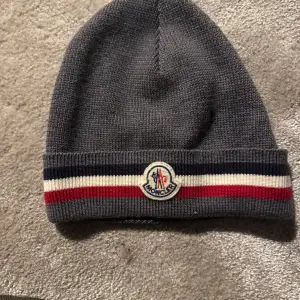 Säljer en snygg grå stickad mössa från Moncler. Den har en klassisk design med Monclers logga framtill och ränder i rött, vitt och blått. Mycket bra skick och sparsamt använd. Pris kan diskuteras🤩