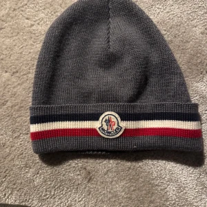 Grå stickad mössa från Moncler - Säljer en snygg grå stickad mössa från Moncler. Den har en klassisk design med Monclers logga framtill och ränder i rött, vitt och blått. Mycket bra skick och sparsamt använd. Pris kan diskuteras🤩