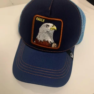 Blå keps med örn broderi - Snygg blå keps med ett coolt broderi av en örn på framsidan. Kepsen har en mesh-baksida i ljusblått för bra ventilation och en justerbar rem för perfekt passform. Perfekt för sommarens äventyr eller bara för att se stilren ut till vardags.