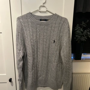 Grå stickad tröja från Polo Ralph Lauren - Säljer en snygg grå stickad tröja från Polo Ralph Lauren i storlek M. Tröjan har ett klassiskt flätmönster och den ikoniska loggan broderad på bröstet. Perfekt för kyliga dagar och passar både till vardags och lite finare tillfällen. Helt ny med tag kvar!