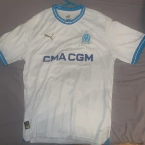 Marseille tröja fotbollströja  - Säljer en vit fotbollströja från Puma med blå detaljer och korta ärmar. Tröjan har en snygg design med klubbens logga och sponsortryck på framsidan. Perfekt för fotbollsfans som vill visa sitt stöd. Tröjan är i mycket bra skick och redo för nya äventyr på planen eller läktaren.