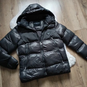 Svart pufferjacka från Alessandro - Säljer en snygg svart pufferjacka från Alessandro Zavetti. Den är perfekt för kalla dagar med sin tjocka vaddering och har en praktisk huva. Jackan har dragkedja framtill och två fickor med dragkedjor. Den är i mycket bra skick och passar perfekt för vintern. Perfekt för dig som vill hålla stilen i kylan! 