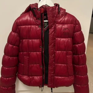 Vinröd pufferjacka - Säljer en snygg vinröd pufferjacka med huva. Den är i mycket bra skick och har en normal passform. Perfekt för höst och vinter!  Nypris 1600kr