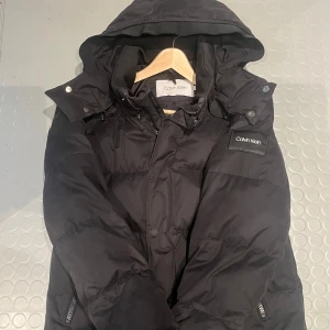 Svart dunjacka Calvin Klein - Säljer en snygg svart dunjacka från Calvin Klein. Den har en praktisk huva och dragkedja framtill. Perfekt för kalla vinterdagar. Jackan är i mycket bra skick och har en stilren design med märkets logga på vänstra armen. Den har en de effekt som är på bild 3💫
