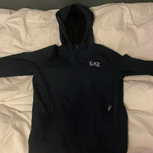 Mörkblå huvtröja från EA7 - Säljer en Mörkblå huvtröja från EA7 i helt okej skick, det finns ett hål på vänstra armen. Den har en dragkedja framtill och är perfekt för höst och vår. Tröjan har långa ärmar och en bekväm passform. Perfekt för både träning och vardag!