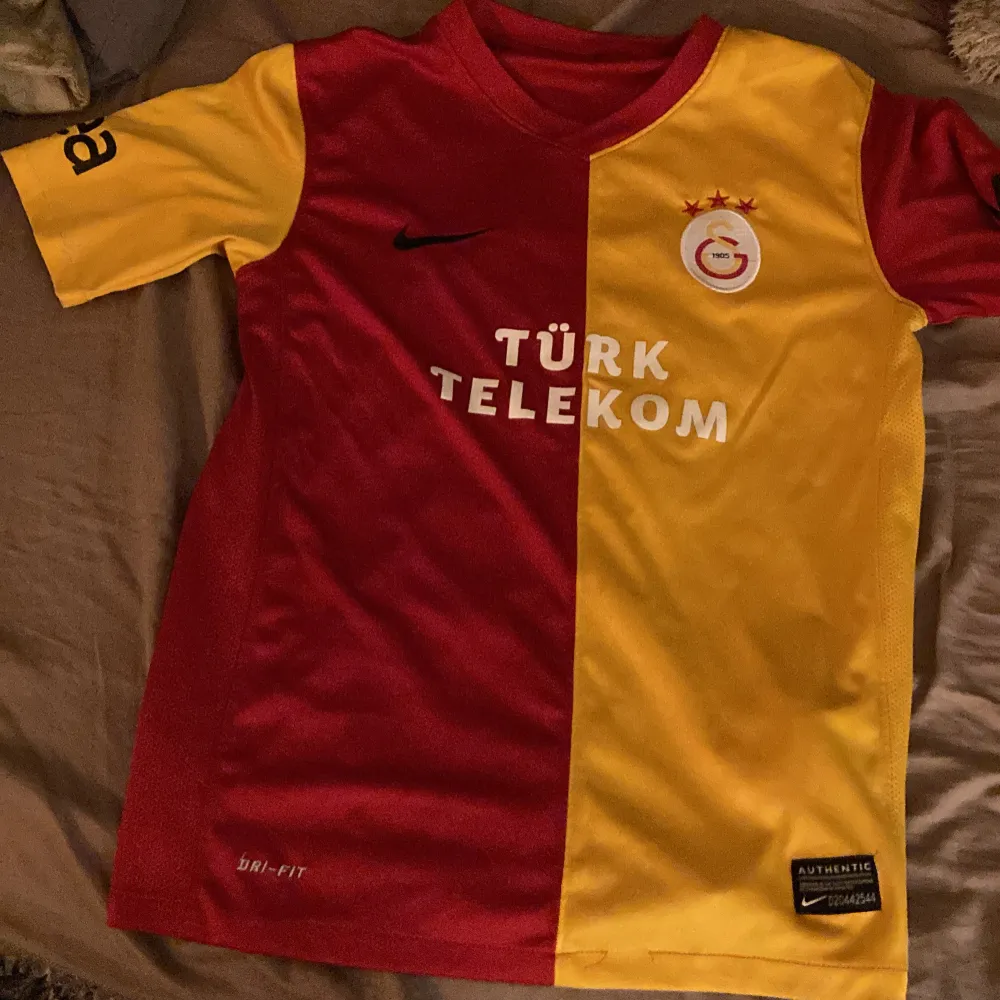 Säljer en Galatasaray fotbollströja från Nike i rött och gult. Den är kortärmad och har sponsorloggor från Türk Telekom och Avea. Tröjan är i mycket bra skick och har en autentisk etikett. Perfekt för fans som vill visa sitt stöd! Passar för 10-12 åringar.. T-shirts.