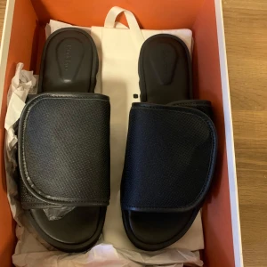 Svarta sandaler från Rodebjer - Snygga svarta sandaler från Rodebjer i nytt skick. De har en bred rem över foten med kardborreband för enkel justering. Sulan är tjock och har ett coolt, tandat mönster för extra grepp. Perfekta för en avslappnad sommarlook eller att glida runt hemma. Kommer i originalförpackning.