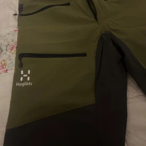 Gröna och svarta byxor från Haglöfs - Säljer ett par gröna och svarta byxor från Haglöfs i mycket bra skick. De har flera praktiska dragkedjefickor och är perfekta för utomhusaktiviteter. Byxorna är tillverkade i slitstarkt material och har en bekväm passform. Perfekta för höst- och vårvandringar!