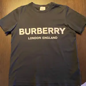 Säljer en snygg svart t-shirt från Burberry med vit text 'BURBERRY LONDON ENGLAND' på framsidan. T-shirten är i mycket bra skick och är inte använd så mycket. Kommer med originaltaggar och förpackning.