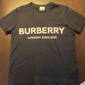 Svart t-shirt från Burberry - Säljer en snygg svart t-shirt från Burberry med vit text 'BURBERRY LONDON ENGLAND' på framsidan. T-shirten är i mycket bra skick och är inte använd så mycket. Kommer med originaltaggar och förpackning.