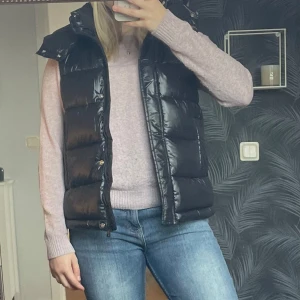 Svart dunväst - Säljer en snygg svart dunväst i glansigt material. Perfekt för höst och vår, med dragkedja och knappar framtill. Västen har en normal passform och är superbekväm att ha på sig över en tröja eller hoodie. Storlek S och är ursprungligen från Mango. Aldrig använd, endast testad! 