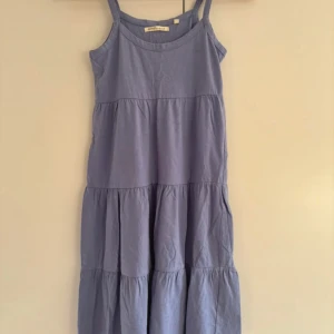 Ljusblå klänning från Emoi - Säljer en söt ljusblå klänning från Emoi i 100% bomull. Perfekt för sommardagar med sin luftiga design och bekväma passform. Klänningen är ärmlös och har en avslappnad stil med flera volanger. Perfekt för både vardag och festliga tillfällen! Köparen betalar frakt. 