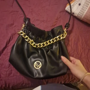 Svart axelväska med guldkedja - Snygg svart axelväska i syntetmaterial med en cool guldkedja som detalj. Väskan har en rund guldfärgad logotyp på framsidan och en praktisk öppning upptill. Perfekt för både vardag och fest! Köpt för 300, säljer för halva priset men det kan diskuteras